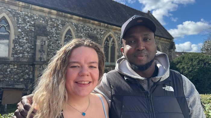 Two people smiling outside an old stone church on a bright, sunny day.