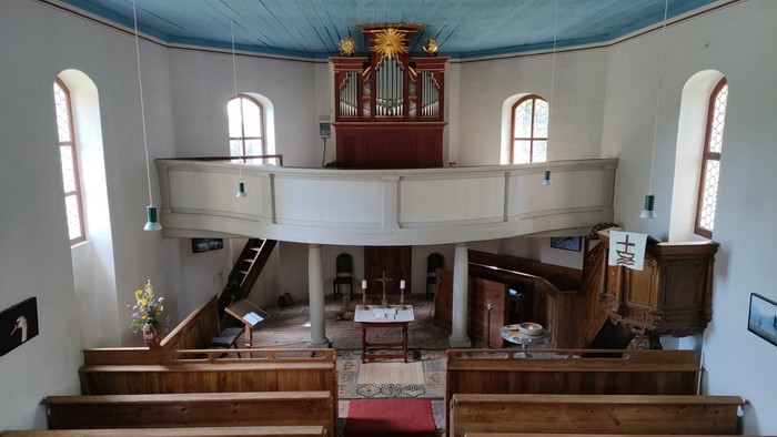 Idyllisches Kircheninneres mit hölzernen Kirchenbänken, einer erhöhten Orgel und einem besinnlichen Altarbereich.