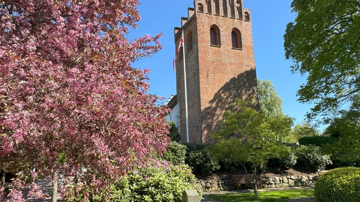 Historisk murstens tårn indrammet af blomstrende træer og frodig grønne omgivelser under en klar blå himmel.