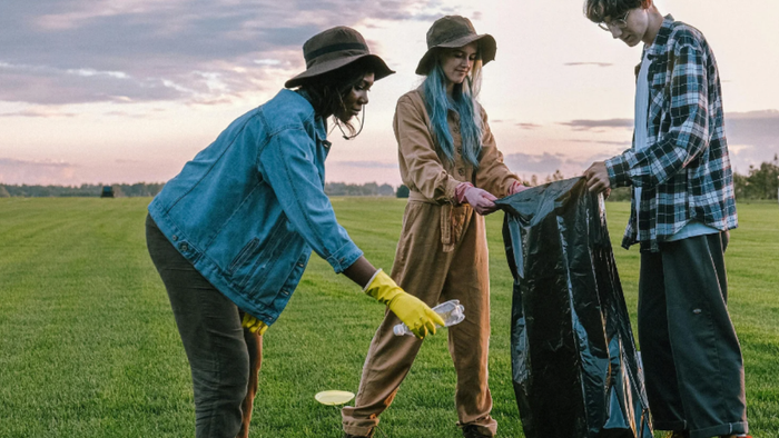 Drei Personen mit Handschuhen sammeln Müll auf einem grünen Feld während des Sonnenuntergangs.