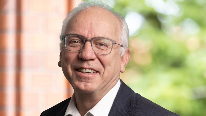 Lächelnder Mann mit Brille, der im Freien ein Sakko und ein weißes Hemd trägt.
