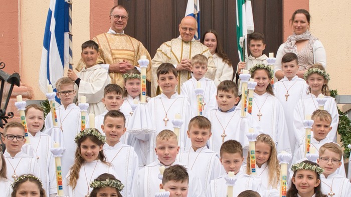 Eine Gruppe von Kindern in weißen Gewändern und Geistlichen steht vor einer Kirche und hält Fahnen und Kerzen.