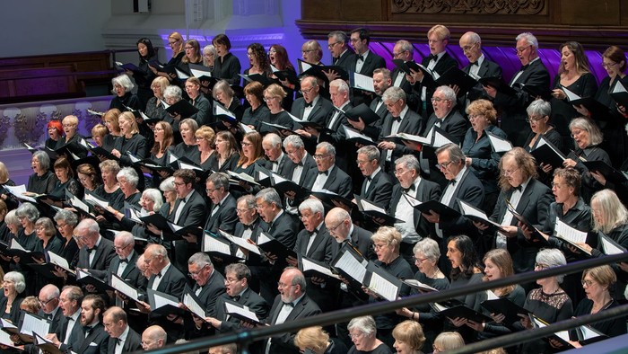 Großer Chor und Orchester, die gemeinsam in einem prächtigen Konzertsaal auftreten.