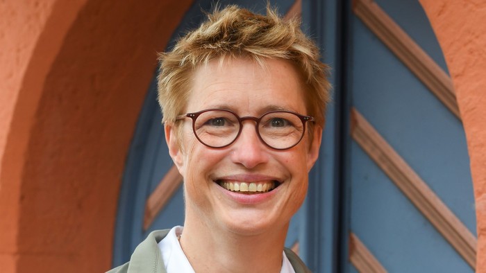 Eine fröhliche Person mit Brille, einem grünen Mantel und einem weißen Oberteil vor farbenfrohen architektonischen Bögen.