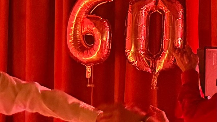 Eine Person, die glänzende Folienballons in der Form der Zahl „60“ vor einem roten Hintergrund hält.