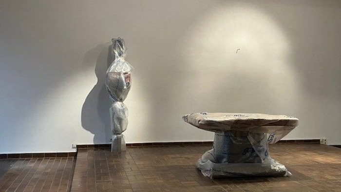 Moderne Kunstinstallation mit drapierten Skulpturen und einem strukturierten Rundtisch in einem minimalistischen Galerieraum.