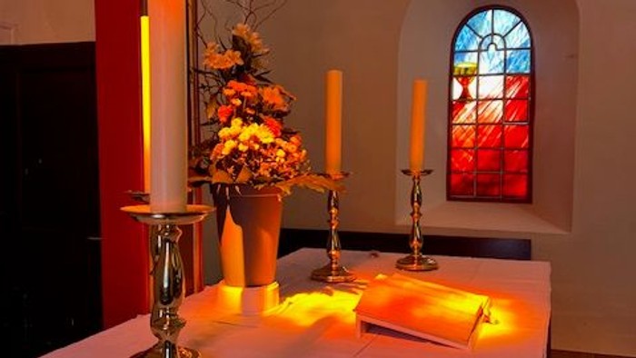Elegante Tischdekoration mit brennenden Kerzen, Blumenarrangement und einem Buntglasfenster in einer ruhigen Innenraumeinrichtung.