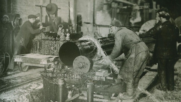 Historisches Schwarz-Weiß-Foto von Arbeitern, die Maschinen in einer geschäftigen Industrieanlage bedienen.