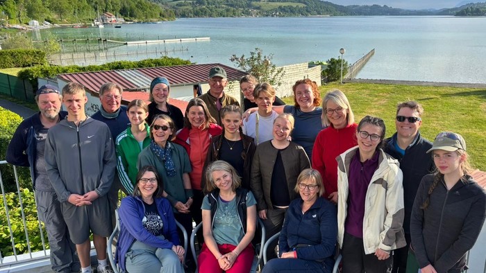 Gruppe posiert gemeinsam an einem malerischen Seeufer mit Bergen im Hintergrund.