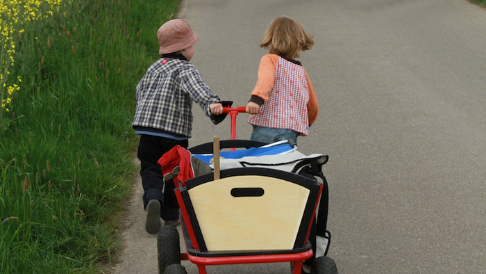 Zwei Kinder schieben einen roten Wagen auf einer Straße.