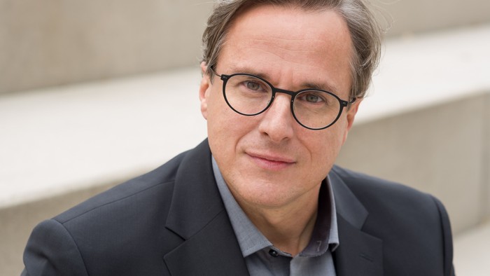 Ein Mann in einem dunklen Anzug und einer Brille posiert im Freien vor einem neutralen Hintergrund.