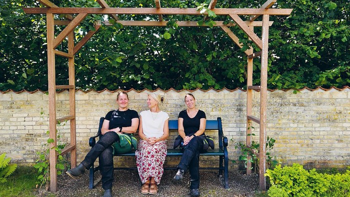 præst Kristine, graver Ida og gravermedhjælper Maria sidder på en bænk under en pergola