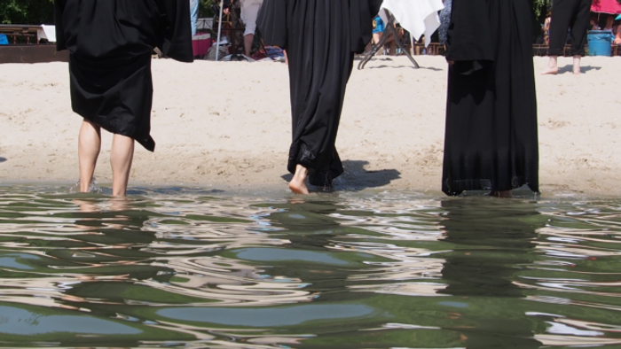 Menschen in langen schwarzen Roben, die durch flaches, spiegelndes Wasser an einem Strand waten.