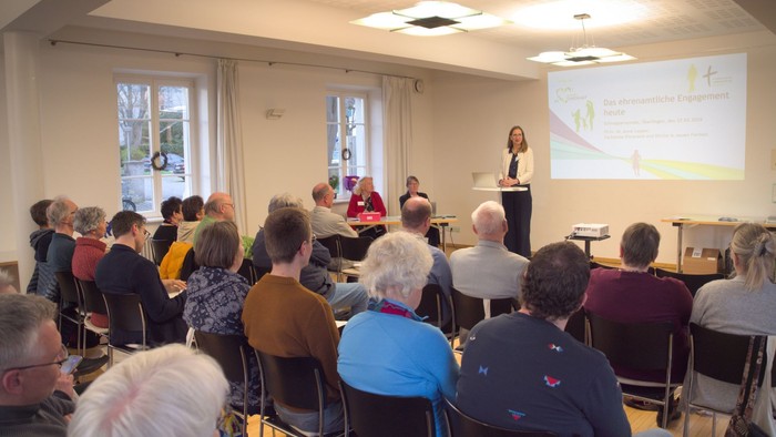 Eine Gruppe von Menschen hört aufmerksam einem Sprecher bei einer Präsentation oder Besprechung in einem Raum mit in Reihen angeordneten Stühlen zu.