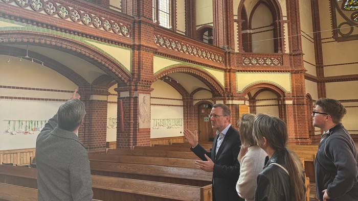 Eine Reisegruppe hört einer Erklärung im Inneren einer prunkvollen, historischen Kirche mit hölzernen Kirchenbänken und Buntglasfenstern zu.