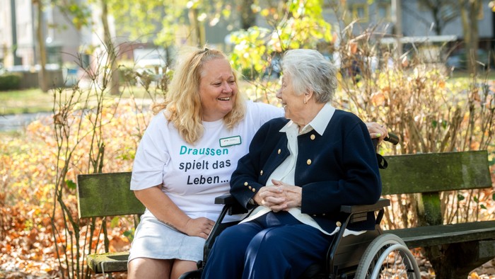 zwei Frauen sitzen auf einer Bank. Eine davon alt und im Rollstuhl. Im Hintergrund ein Park