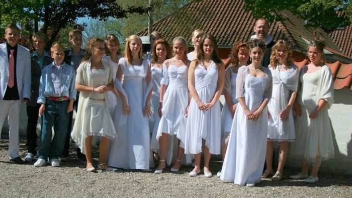 En gruppe børn i hvide tøjer står foran et hus med røde tag.