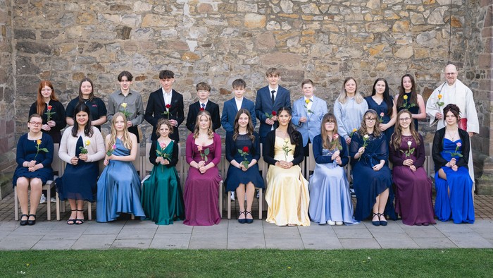 Offizielles Gruppenfoto junger Männer und Frauen in eleganten Kleidern und Anzügen vor steinernem Hintergrund.