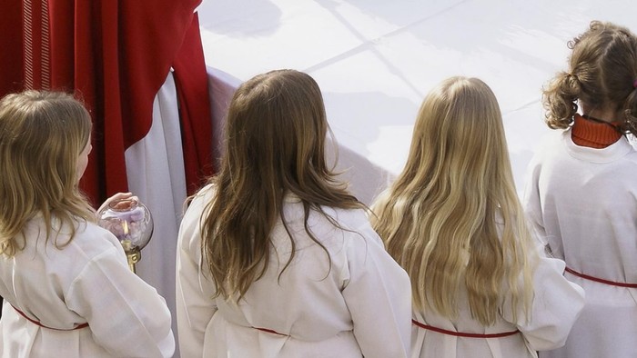 Vier junge Mädchen in weißen Gewändern mit roten Schärpen stehen einem religiösen Altar zugewandt.