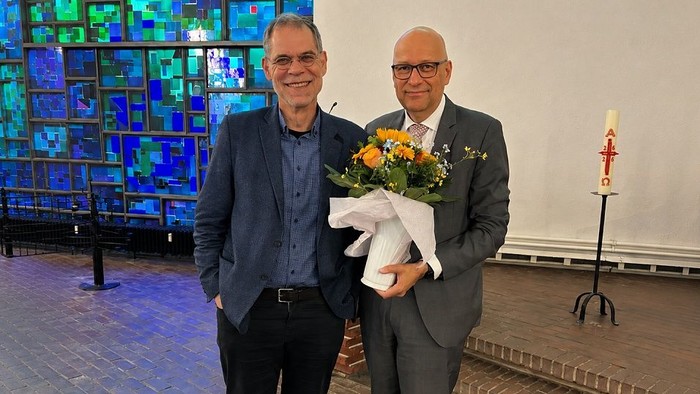 Zwei Männer stehen in einer Kirche vor bunten Fenstern. Einer hält Blumen. Beide lächeln freundlich in die Kamera hinein heute.