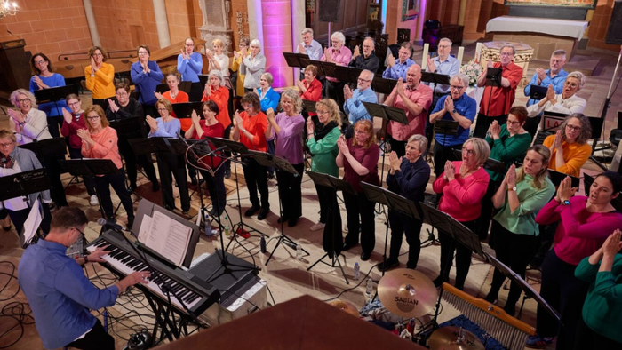 Eine vielfältige Gruppe von Sängerinnen und Sängern und ein Pianist, die gemeinsam in einem großen Innenraum auftreten.