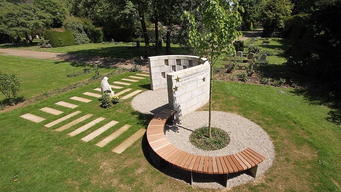Ein Denkmal aus einer halbrunden Mauer und einer halbrunden Bank auf einem Friedhof, aus der Luft gesehen.