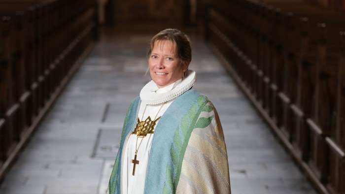 En person i religiøst embedstøj står og smiler i midtergangen i en stor, svagt oplyst kirke.