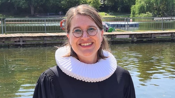 Eine Frau in Abschlusskleidung steht am Flussufer und lächelt.