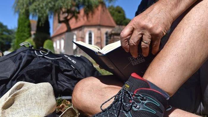 Eine Person sitzt mit Wanderkleidung und Rucksack auf einer Wiese und liest in einer kleinen Bibel. Im Vordergrund sind Wanderschuhe und Gepäck zu sehen, im Hintergrund eine Kirche unter blauem Himmel. Die Szene vermittelt eine ruhige Pause beim Pilgern o