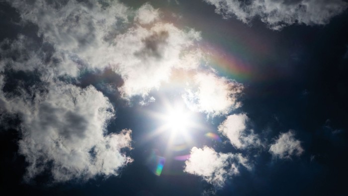 Sonne, die aus den Wolken hervorbricht. In der Mitte ein Sonnenstern und um die Sonne herum ein Lichterkranz. Die Spektralfarben am Himmel erscheinen um die Sonne herum. Farbaufnahme im Gegenlicht.