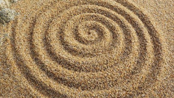 Nahaufnahme einer spiralförmig in feinen Sand gezeichneten Linie. Die konzentrischen Kreise erinnern an ein Labyrinth oder Meditationssymbol und vermitteln Ruhe, Achtsamkeit und innere Sammlung.