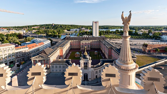 Das Bild zeigt eine Stadtlandschaft mit einem weißen Denkmal und einem Brunnen im Vordergrund.