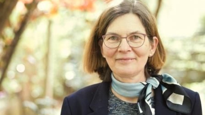 Eine Frau mit Brille und Halstuch posiert vor einem unscharfen Hintergrund aus Bäumen.
