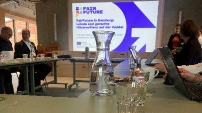 Ausblick von einem Tisch bei der Konferenz. Zu sehen sind Notizenbücher, Gläser und ein Leinwand mit Präsentation