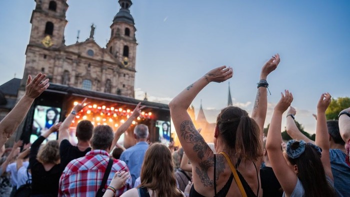 Menge jubelt bei einem Open-Air-Konzert mit historischen Kirchengebäuden im Hintergrund.