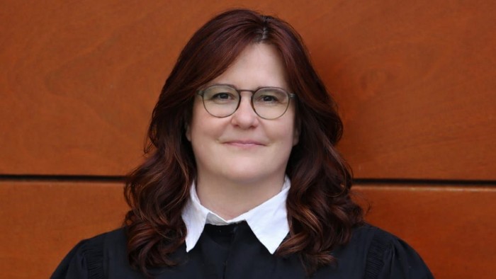 Person in schwarzer Robe mit Brille, die vor einem orangefarbenen Hintergrund lächelt.