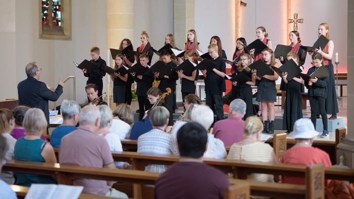 Ein Jugendchor mit Streichinstrumenten tritt in einer Kirche auf, dirigiert von einem Dirigenten, während ein Publikum zuschaut.
