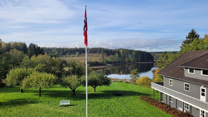 Malerisches Haus am See mit Fahnenmast, üppigem Grün und bewaldeten Hügeln im Hintergrund.
