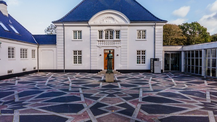 Elegant hvid bygning med blåt tag og et mønstret gårdanlæg med en statue og geometrisk belægning.