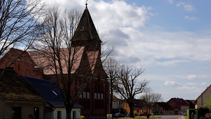 Idyllisches Dorfbild mit einer Kirche mit hohem Kirchturm und Wohnhäusern entlang einer ruhigen Straße.