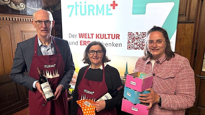 Dr. Jan-Dirk Verwey, Pastorin Susanne Reich und Bildungsreferentin Katharina Schneider halten Wein in den Händen und Schubladenboxen. Sie stehen vor einer beleuchten 7Türme+-Wand in der Brauerkapelle in St. Jakobi.