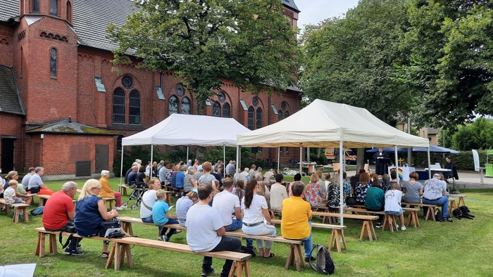 Draußen versammeln sich Menschen unter Zelten in der Nähe einer Backsteinkirche und genießen eine gemeinschaftliche Veranstaltung.
