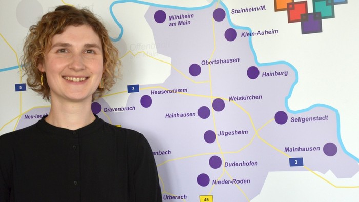 Eine Person steht neben einer bunten Karte, die verschiedene Städte und Orte in einer regionalen Gegend hervorhebt.