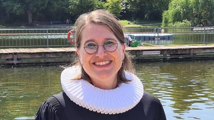 Eine Frau in Abschlusskleidung steht am Flussufer und lächelt.