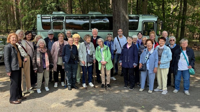 Gruppe älterer Menschen steht lächelnd vor einem Reisebus am Waldrand unter hohen Bäumen.