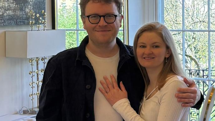 A joyful couple posing together indoors near a window, both wearing casual yet stylish attire and smiling.