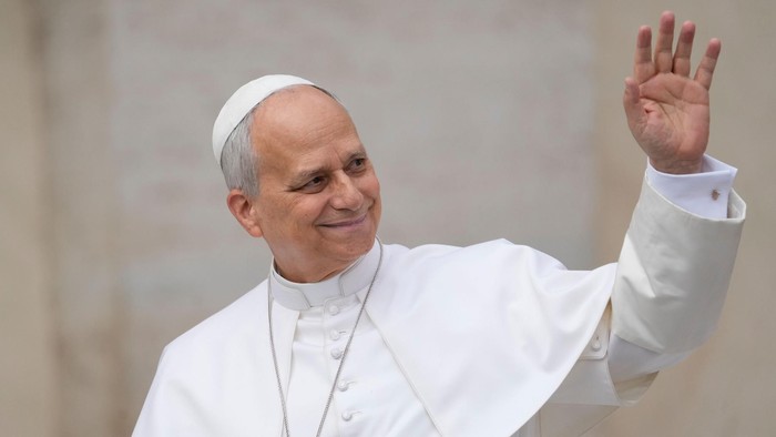 Papst Leo XIV. winkt freundlich Richtung links. Er trägt ein helles Gewand und eine helle Kappe. Der Hintergrund ist heller Stein.