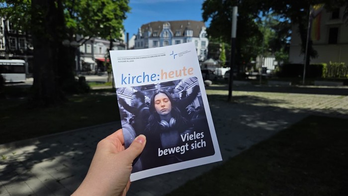 Eine Hand hält eine Zeitschrift mit dem Titel *„kirche:heute“* und der Schlagzeile *„Vieles bewegt sich“* in einer städtischen Umgebung im Freien.