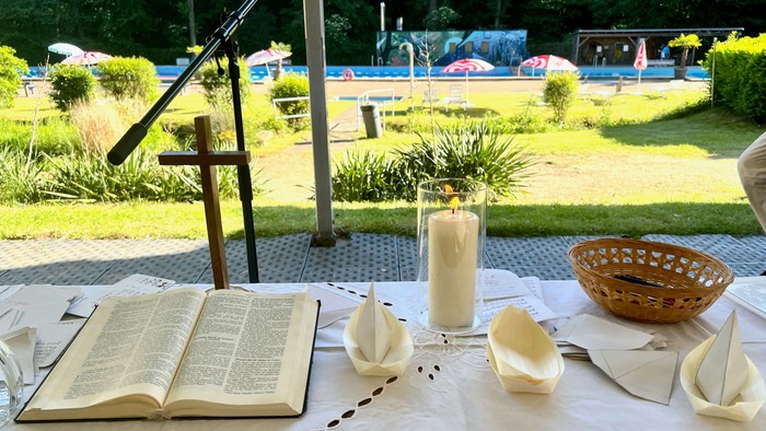 Auf dem Tisch, der als Altar dient, sind eine Bibel, ein Kreuz, ein Windlicht und einige kleine selbstgebastelte Schiffchen zu sehen; im Hintergrund Liegewiese und Schwimmbecken
