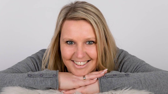Eine Frau mit blondem Haar lächelt in die Kamera und stützt ihr Kinn auf die Hände.